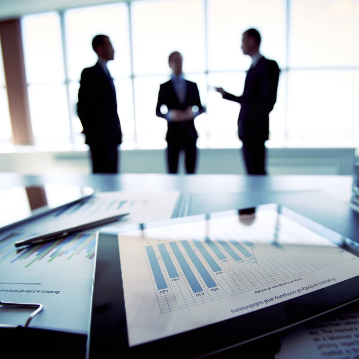 Close-shot of a tablet computer displaying financial data, three businessmen standing in the background