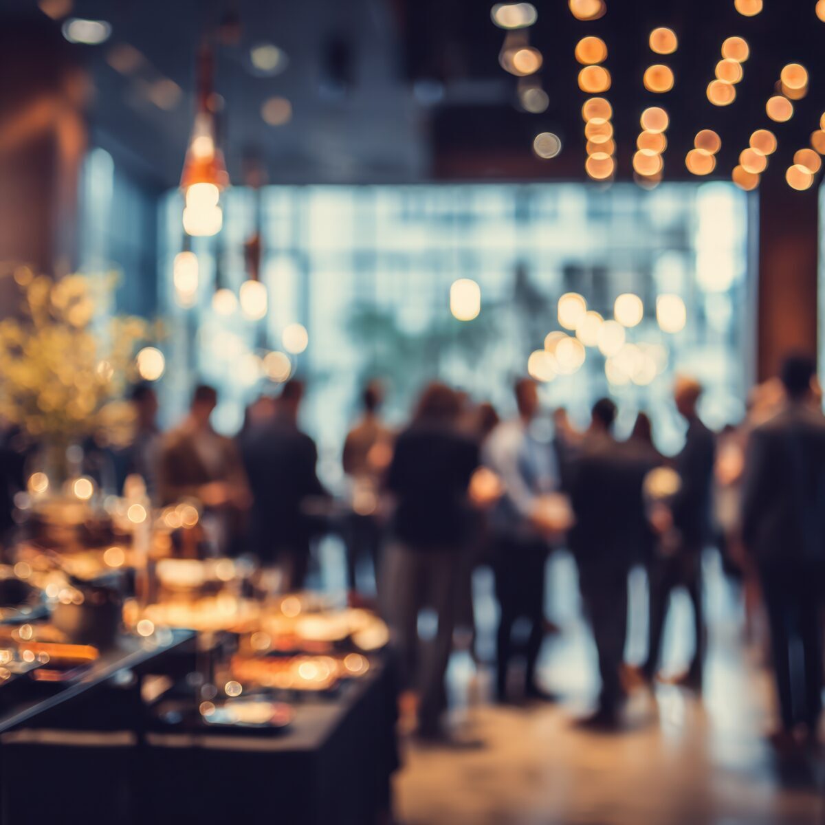 A blurred scene captures a sophisticated social gathering with people mingling near a buffet table under ambient lighting, creating a warm and inviting atmosphere.