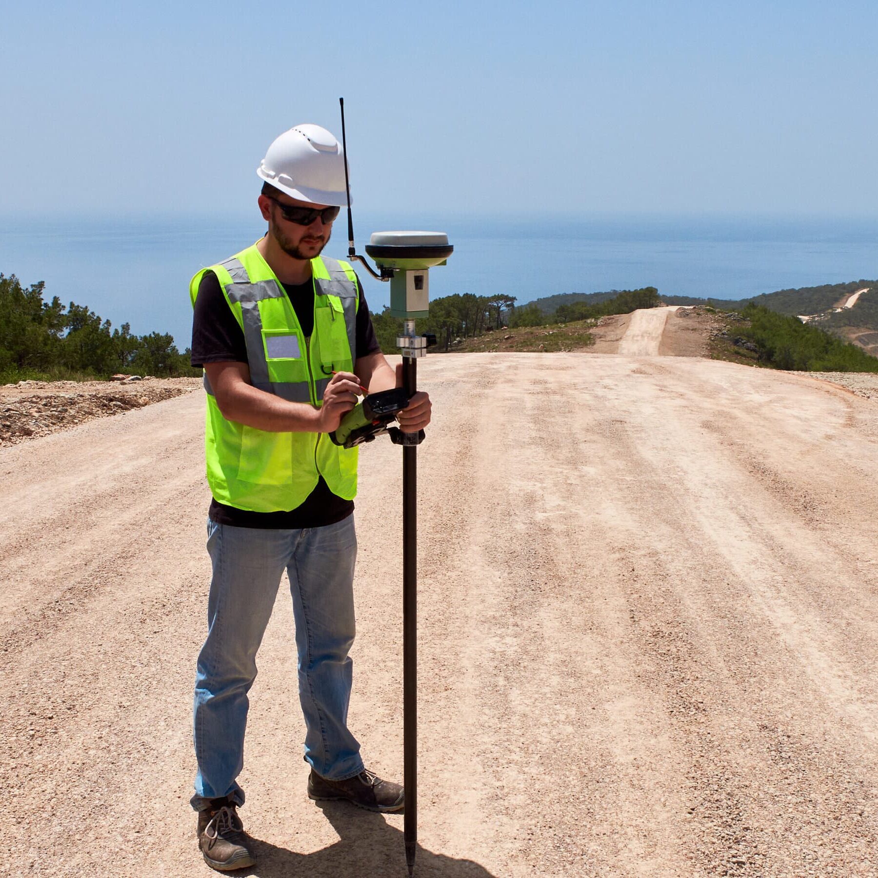 Geodetic engineer surveyor in white hard hat doing measurements with GNSS satellite receiver during road construction works.