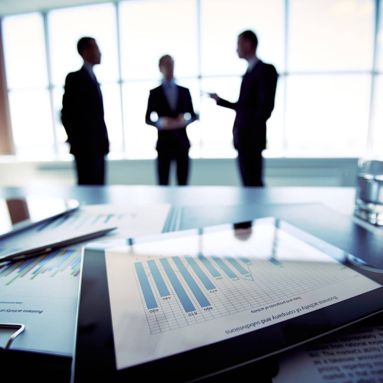 Close-shot of a tablet computer displaying financial data, three businessmen standing in the background