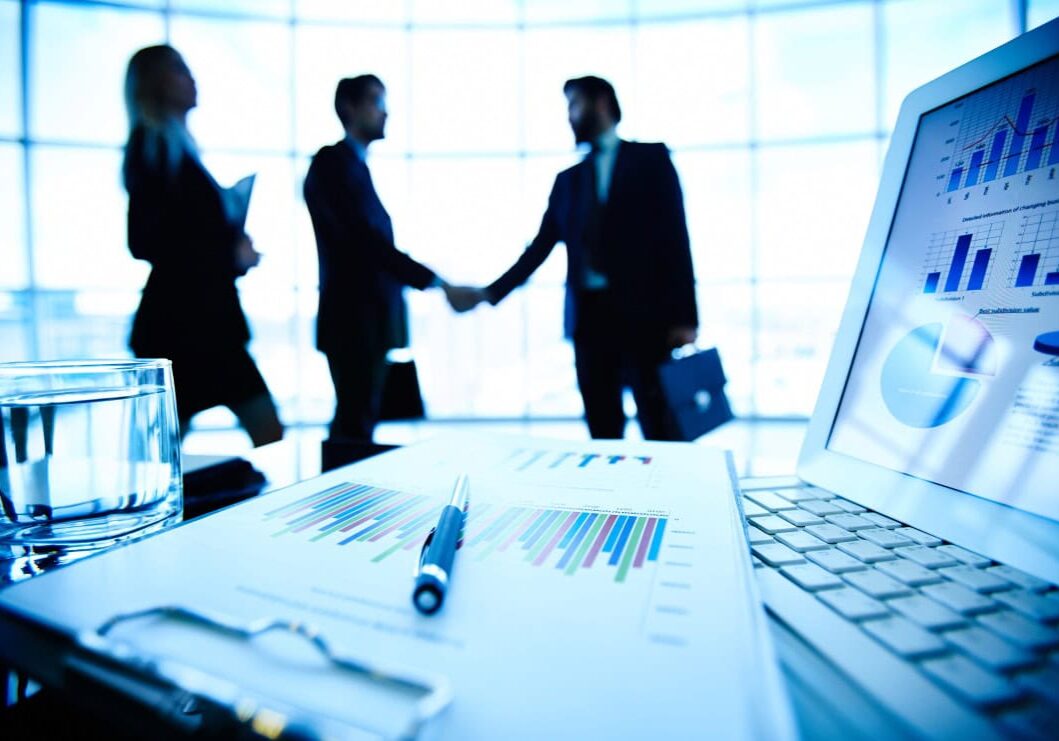 Laptop, financial document with pen and glass of water at workplace on background of three business partners making a deal